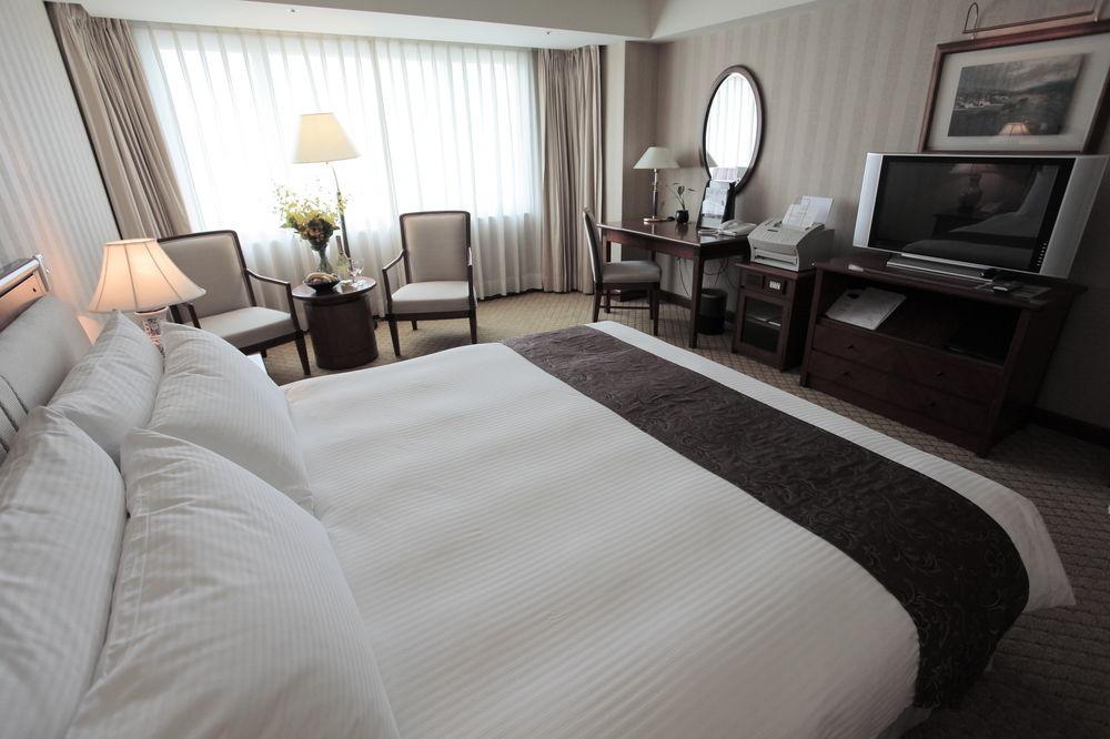 Evergreen Laurel Hotel - Taichung Exterior foto The photo shows a well-furnished hotel room. In the foreground, there is a large bed with white bedding and a dark-colored throw at the foot. Beside the bed, there is a bedside lamp. 

In the background, natural light filters through large windows co