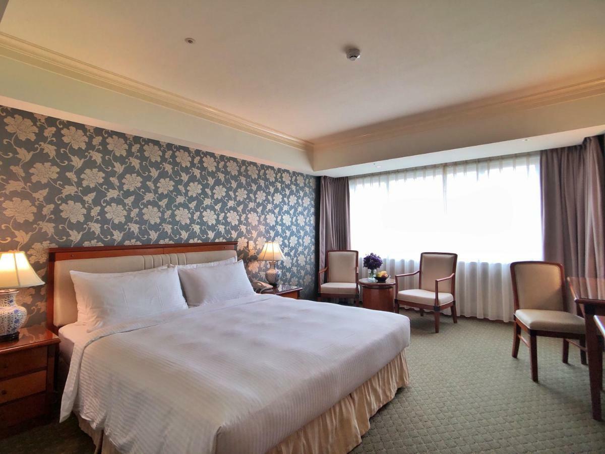 Evergreen Laurel Hotel - Taichung Exterior foto The photo shows a hotel room that features a large bed with a white bedspread positioned against a decorative floral wallpaper. There are two bedside lamps on wooden nightstands flanking the bed. To the side of the room, there are two chairs and a sm