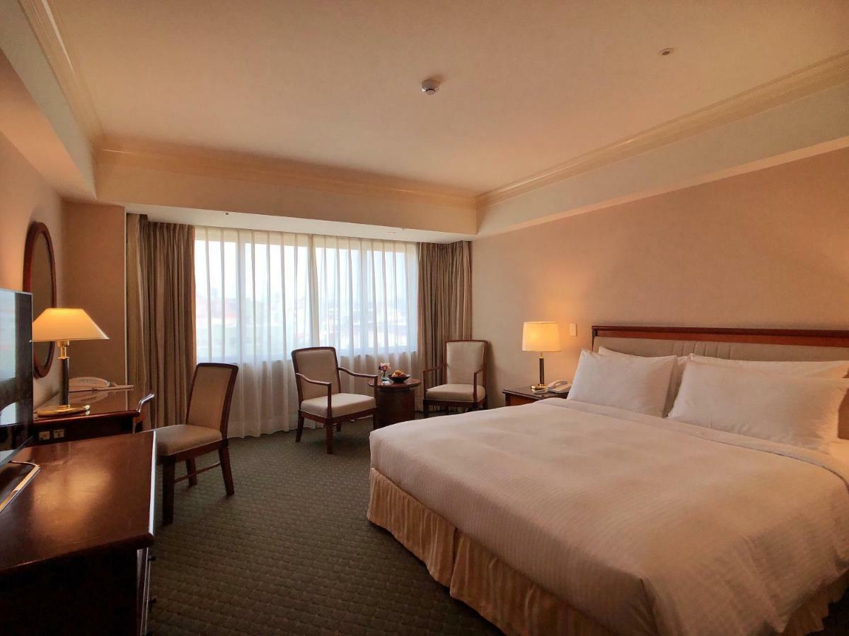 Evergreen Laurel Hotel - Taichung Exterior foto The photo shows a hotel room featuring a large bed with white linens, a wooden bedside table with a lamp, and a flat-screen television. There are two chairs with a small table next to them positioned by a large window, which has sheer curtains lettin